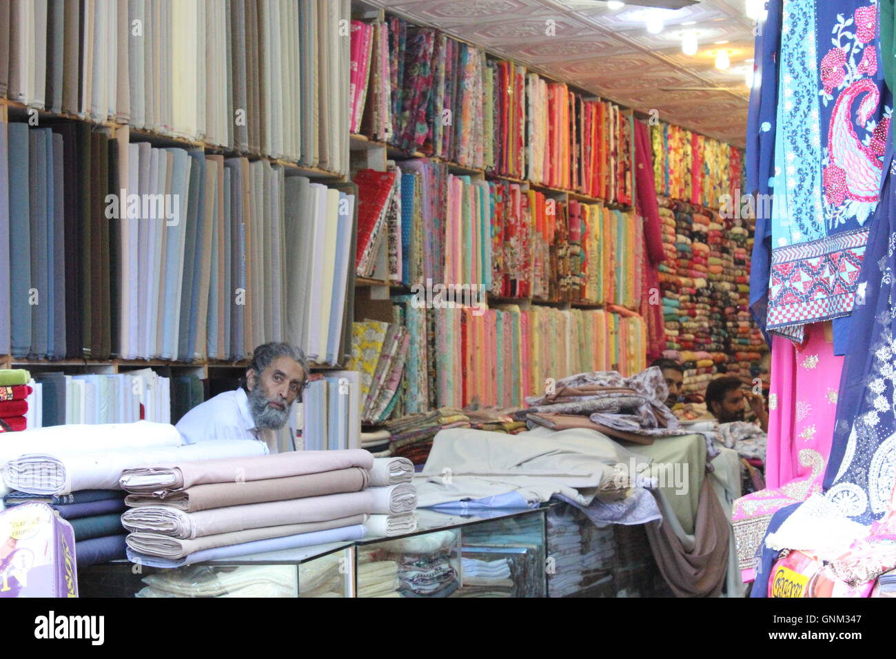 Ein Shop-Betreiber von einem Tuch Shop sitzt an der Theke, zeigt der Hintergrund einen Wand voller bunter als auch monochrome Tücher. Stockfoto