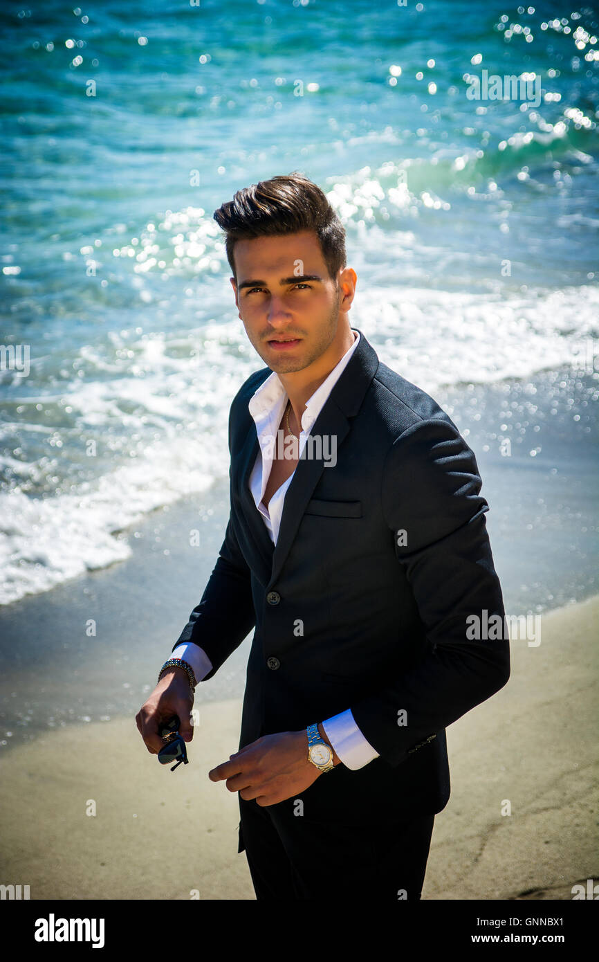 Schönen jungen Mann im klassischen Anzug am Strand Sonnenbrillen beim Blick in die Kamera halten. Wellen des Meeres im Hintergrund Stockfoto