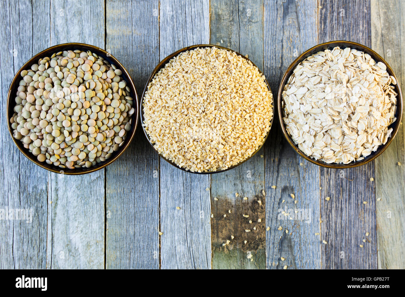Schalen mit Linsen, Bulgur und Haferflocken in einer Reihe auf einem alten Holztisch von oben gesehen. Stockfoto