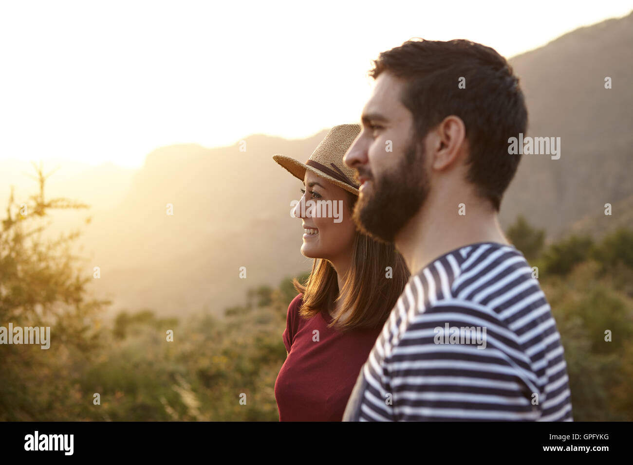 Junges Paar auf der Suche umgeben von Büschen und sehr helle Sonne auf ihrer linken Seite tragen T-shirts und einen Strohhut bei Sonnenuntergang Stockfoto