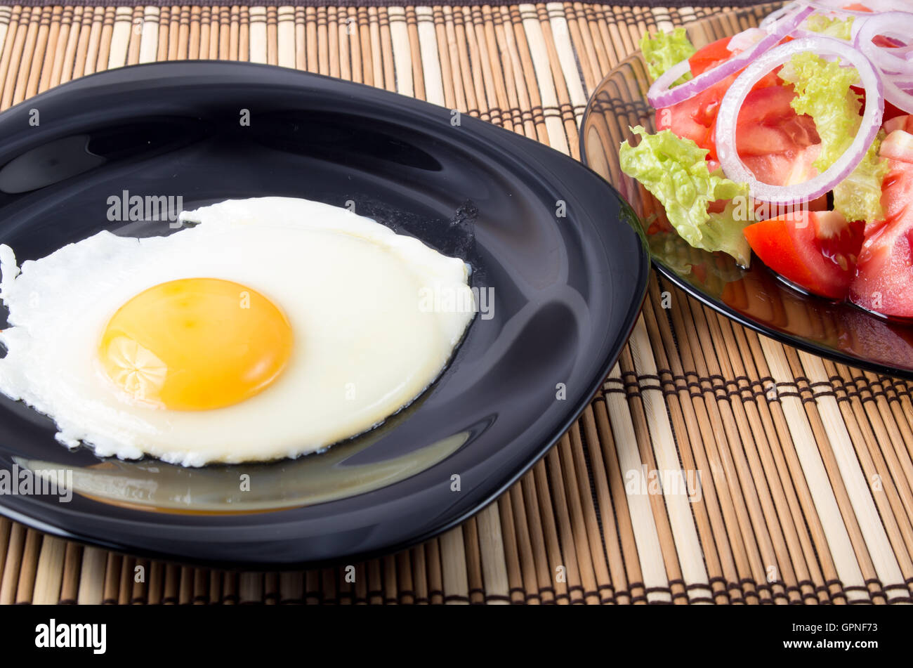 Detailansicht auf Spiegeleier mit Eigelb auf einer schwarzen Platte und einem Salat aus Tomaten, Salat und Zwiebeln auf einem gestreiften Matte Stockfoto