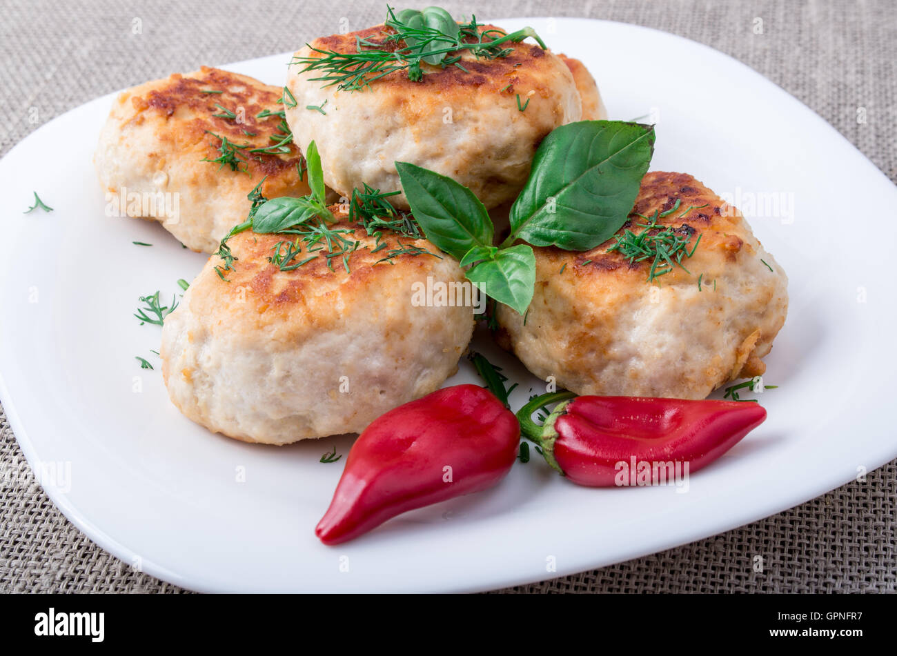 Nahaufnahme auf Frikadelle aus gehacktem Hühnerfleisch auf einem weißen Teller mit rotem Pfeffer und Basilikum auf Sackleinen Hintergrund Stockfoto