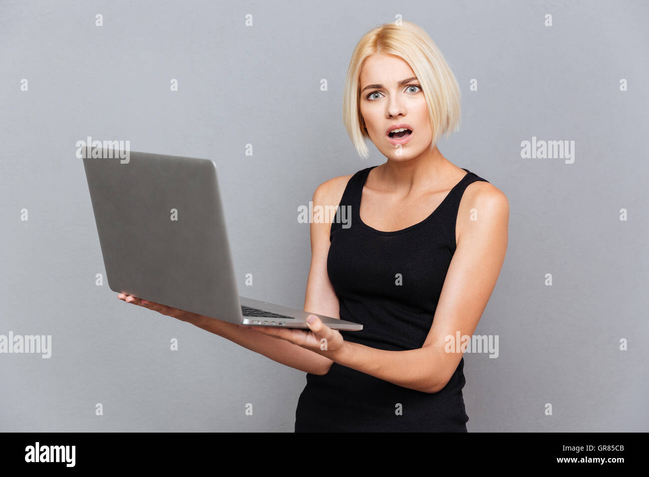 Unglückliche junge Frau mit Laptop über grauen Hintergrund erstaunt Stockfoto