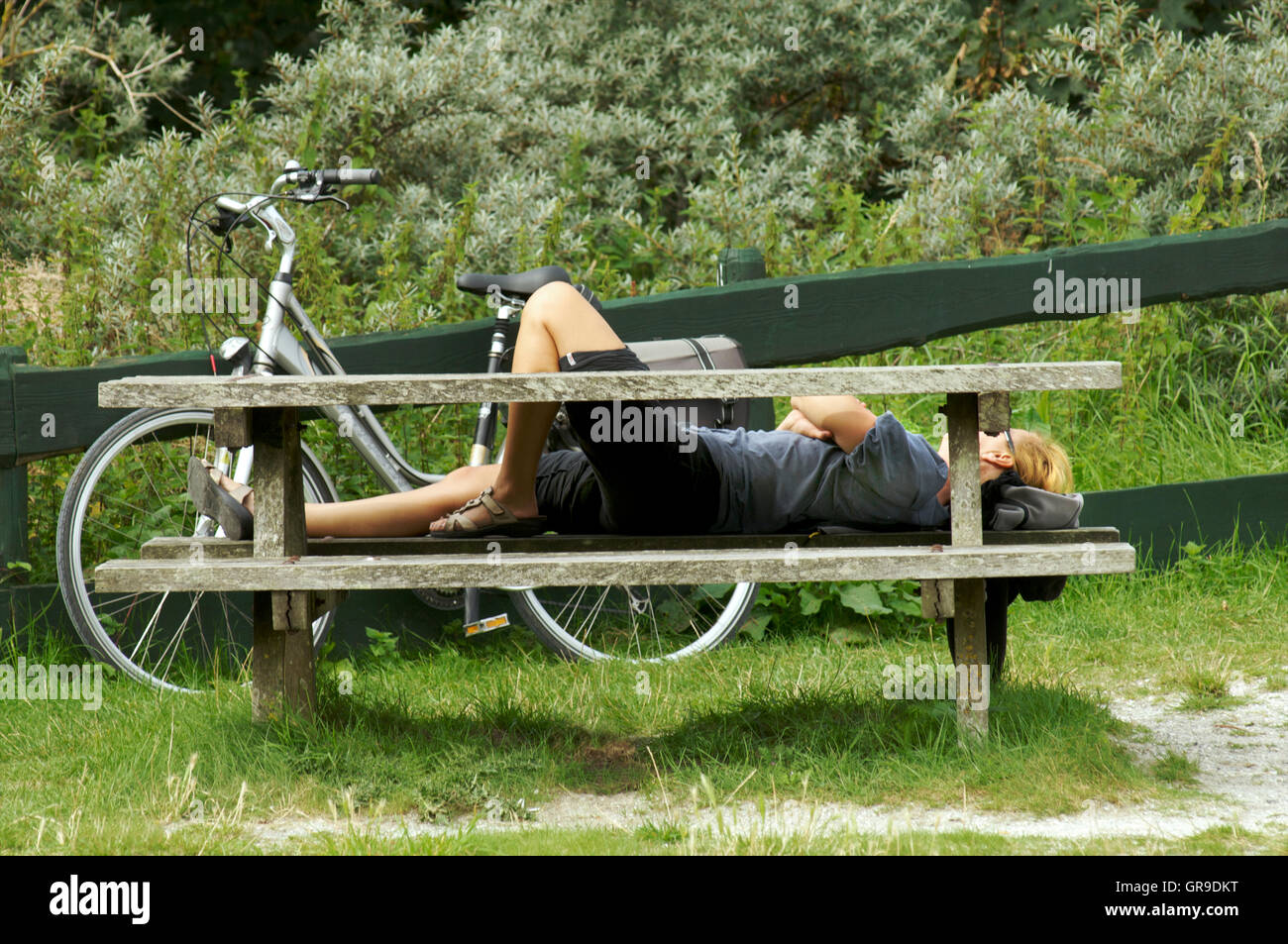 Frau ruht auf einer Holzbank mit ihrem Fahrrad neben ihr während einer Radtour auf der Insel Schiermonnikoog, Niederlande Stockfoto