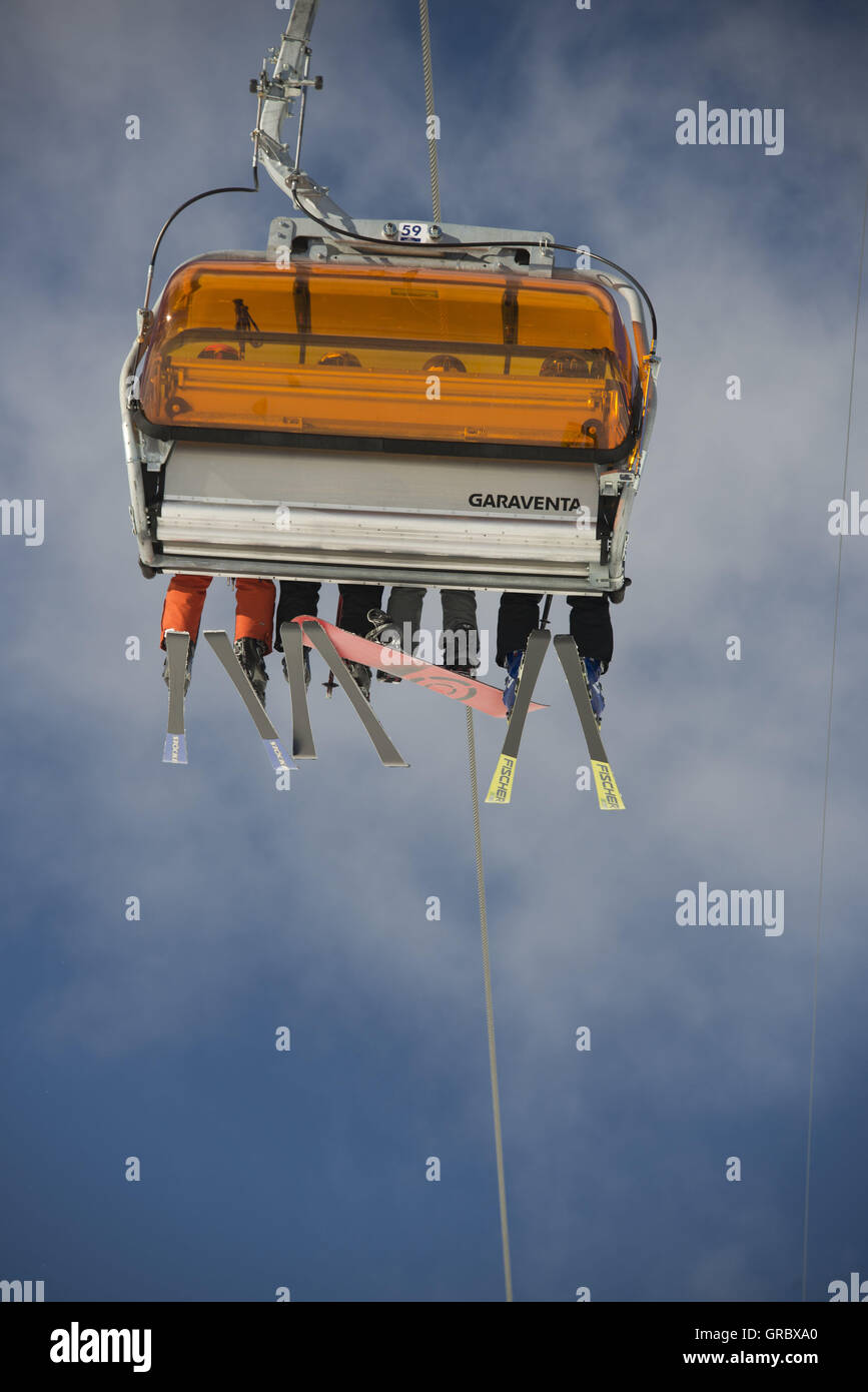 Einzelstuhl aus Sesselbahn mit Orangen Wetterschutzhauben, Beine mit Skiern gegen den blauen Himmel und weiße Wolken von hinten gesehen Stockfoto