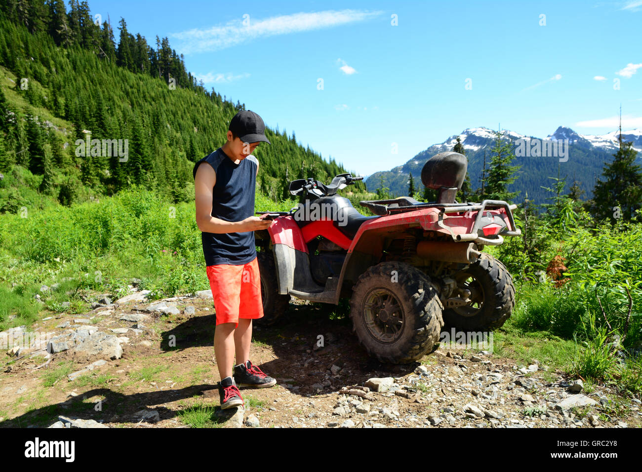 GenY Männchen steht vor Natur Valley neben ATV mit iPhone6 Smartphones Onroute über Kies Trail in der Nähe von Whistler, BC Stockfoto