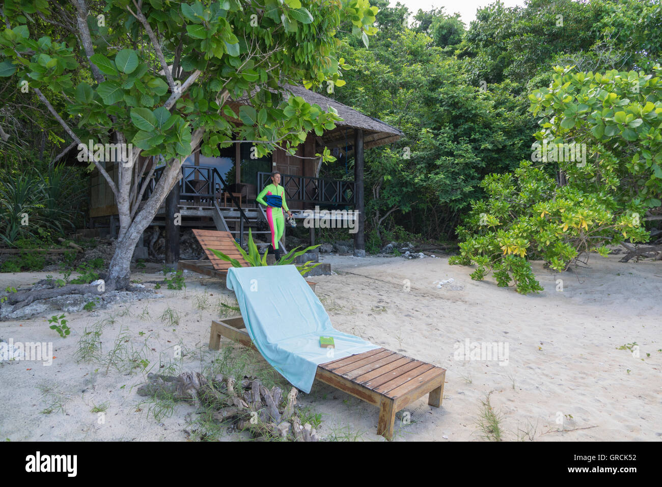 Weibliche Schnorchler vor Holzbungalow, umgeben von üppigen Buschwerk, In den Vordergrund Liegestuhl am Sandstrand Stockfoto