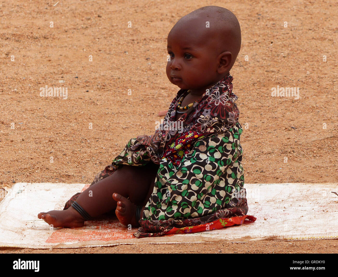Himba-Kind In bunten Kleidern sitzen auf einer Matte Stockfoto