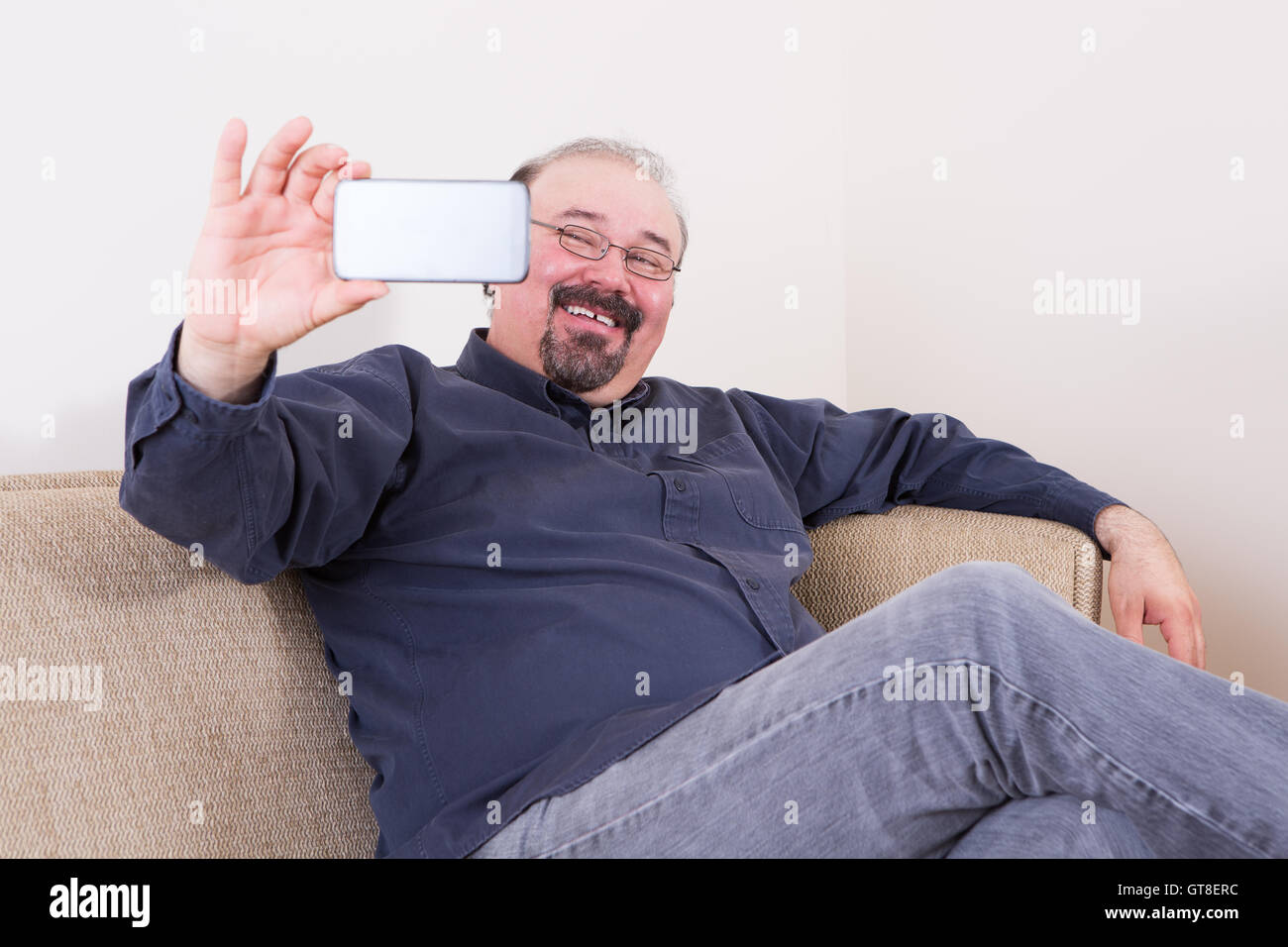 Glücklicher Mann unter einem Selfie für seine Freunde in den sozialen Netzwerken auf seinem Smartphone posiert auf der Couch zu Hause schaut in die Kamera Stockfoto