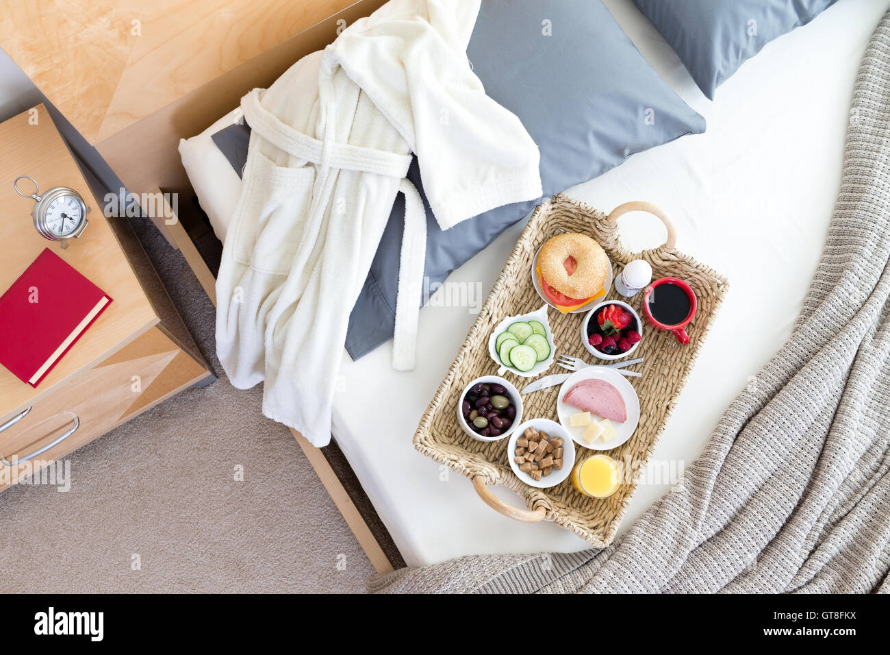 High Angle View of Bademantel und Frühstückstablett auf ungemachten Bett neben Nachttisch mit roten Buch im Hotelzimmer Stockfoto