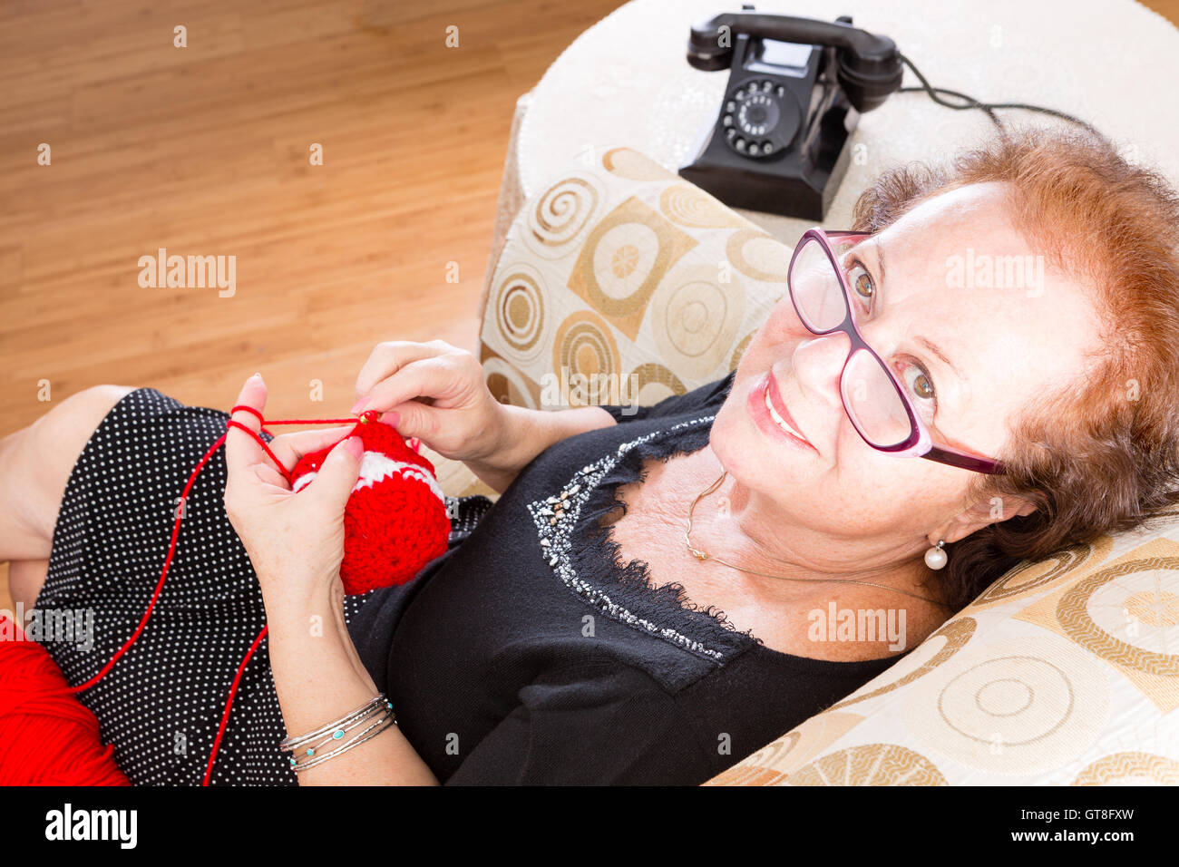 Ältere Großmutter tragen Brillen sitzen auf einem Stuhl nach oben auf die Kamera mit einem Lächeln, old fashioned rotary t stricken Stockfoto