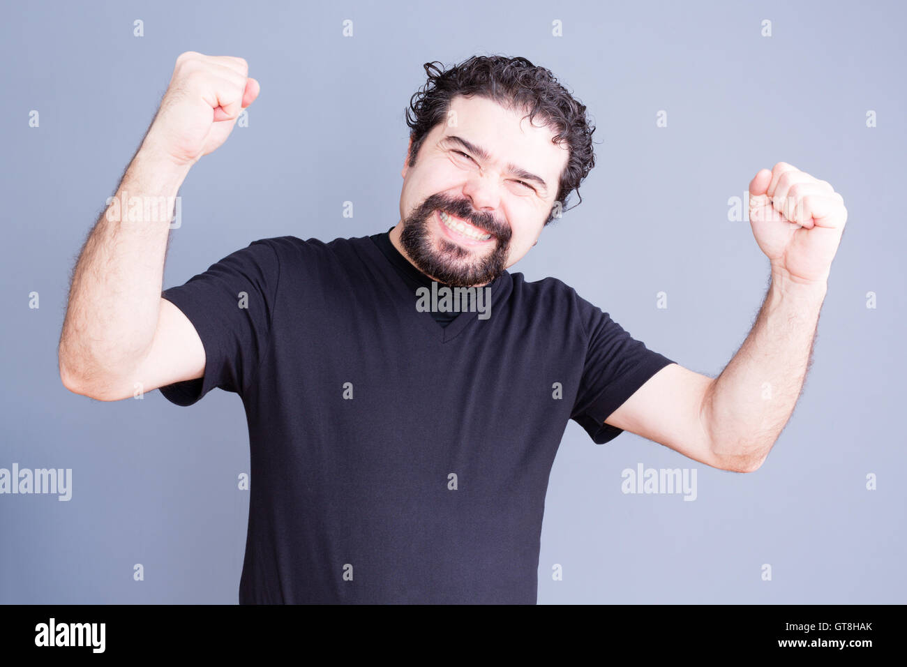 Ein hübscher bärtiger Mann im schwarzen Hemd hochhalten Fäuste mit beschwingt Ausdruck über grauen Hintergrund Stockfoto