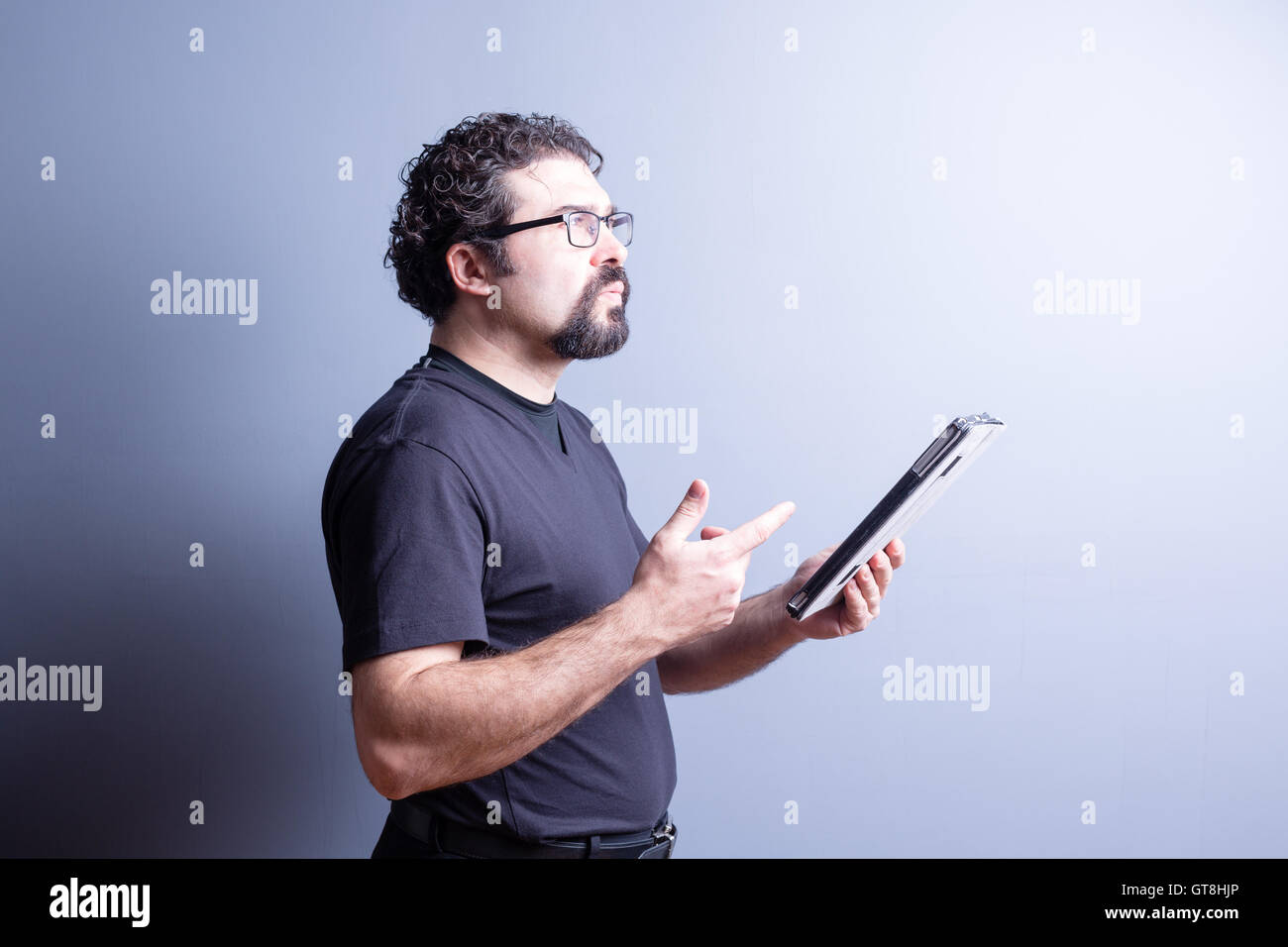 Profil von Mann mit T-Shirt und Brillen tief in Gedanken halten Sie Computer Tablet im Studio mit grauem Hintergrund, Seite Stockfoto