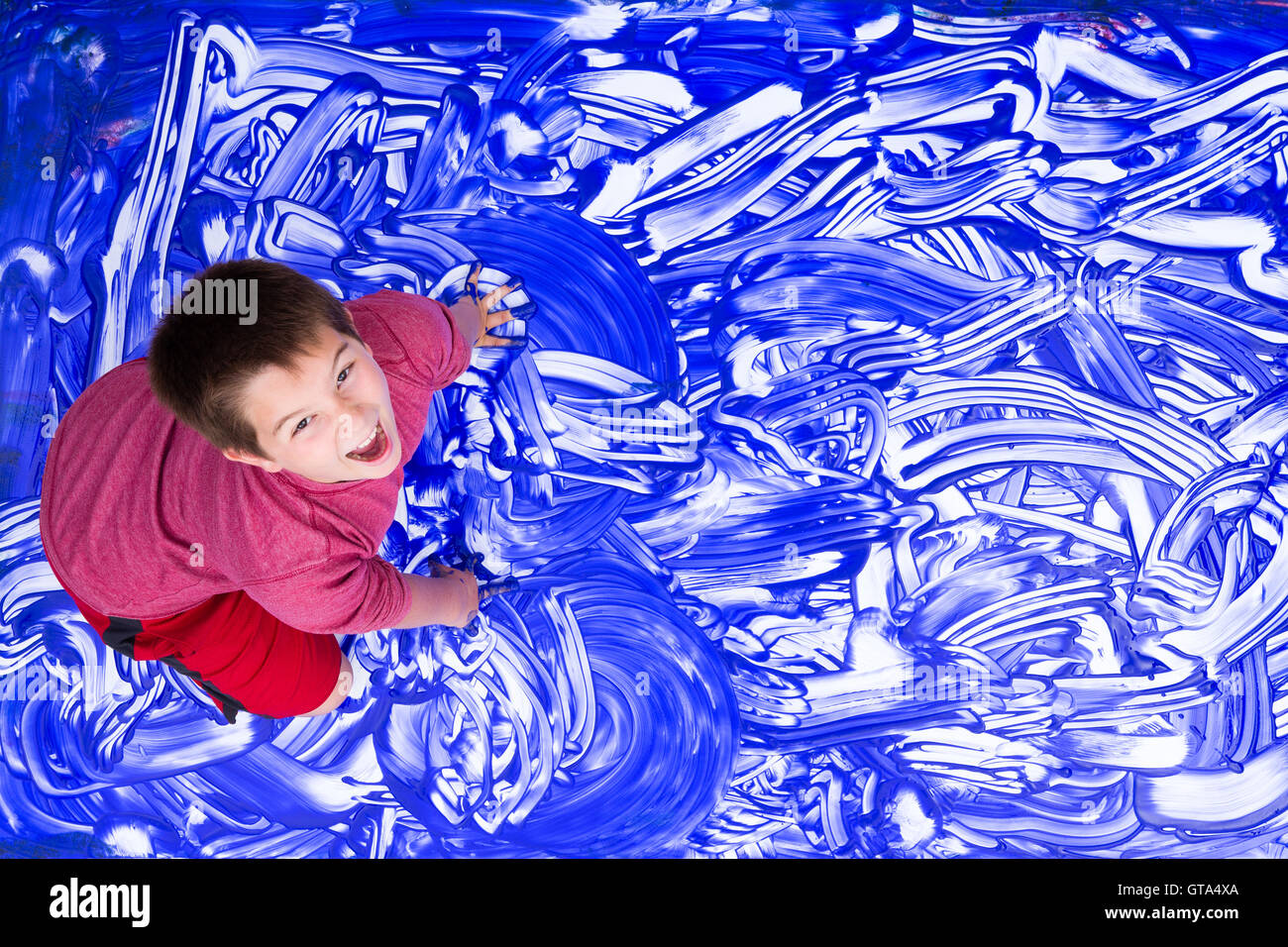 Niedlicher einziger lachender Junge im roten Hemd plantschen Hände in blauer Farbe für abstrakte Wandbild Stockfoto
