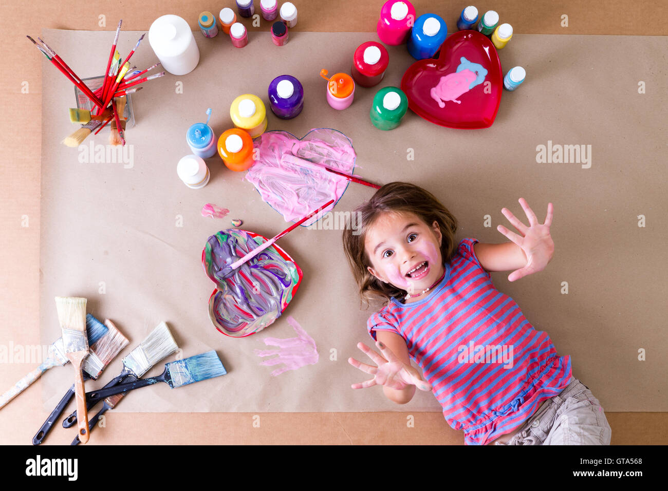 Verspieltes kleines Mädchen umgeben von ihrer Gläser mit bunten Wasserfarben und Pinsel liegen auf einem Blatt braun nachschlagen ein Stockfoto