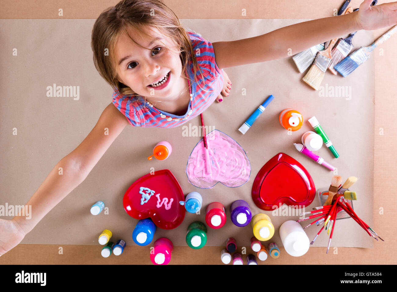 Glückliches kreative kleine Mädchen mit Farben und Pinsel, stehend nach oben auf die Kamera mit ihren ausgestreckten Armen und einem beamin Stockfoto