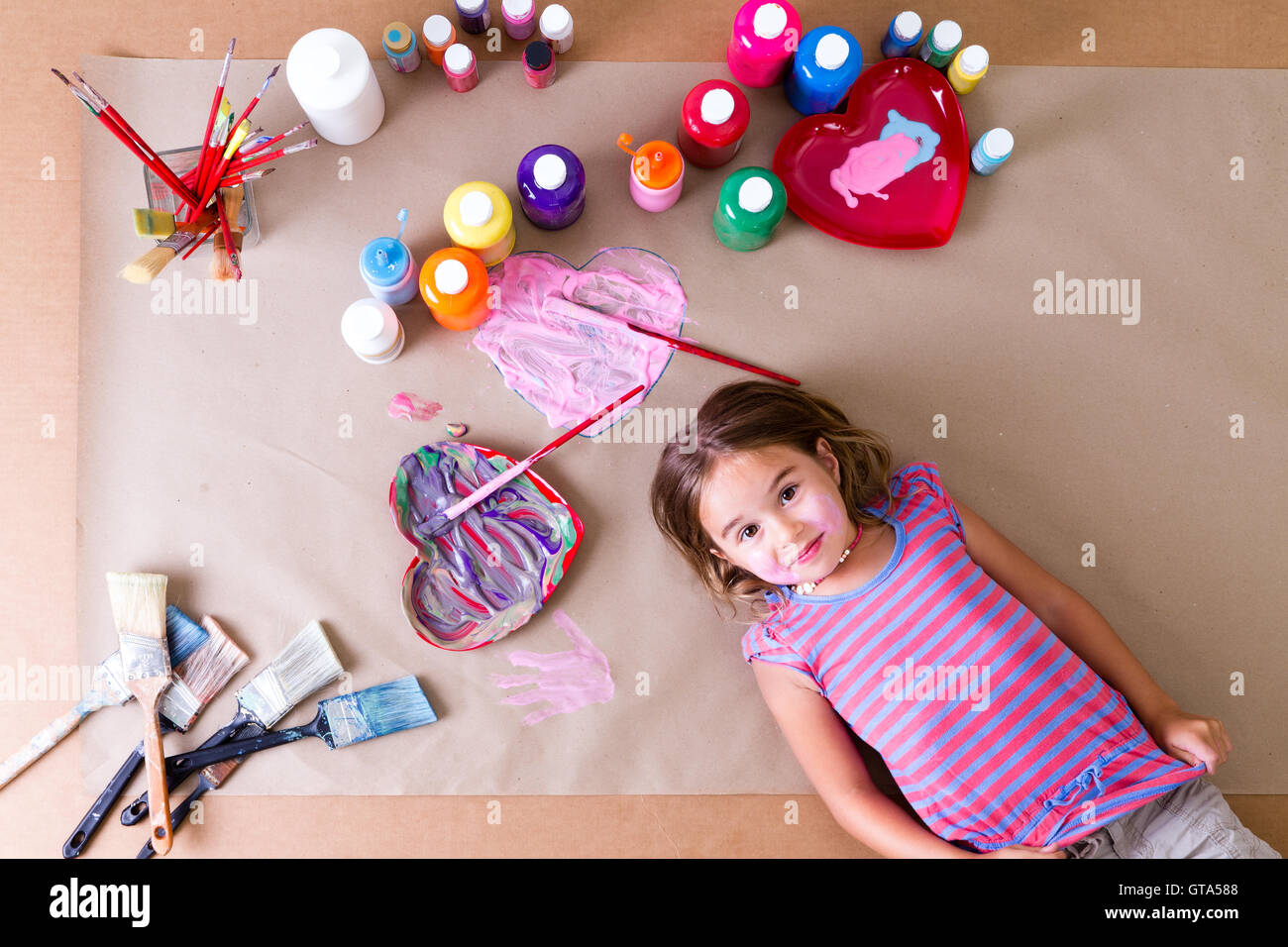Nette hübsche kleine Mädchen Künstler umgeben mit bunten Farben, Pinsel und künstlerische Herz-Design liegen auf einem Blatt braun pape Stockfoto