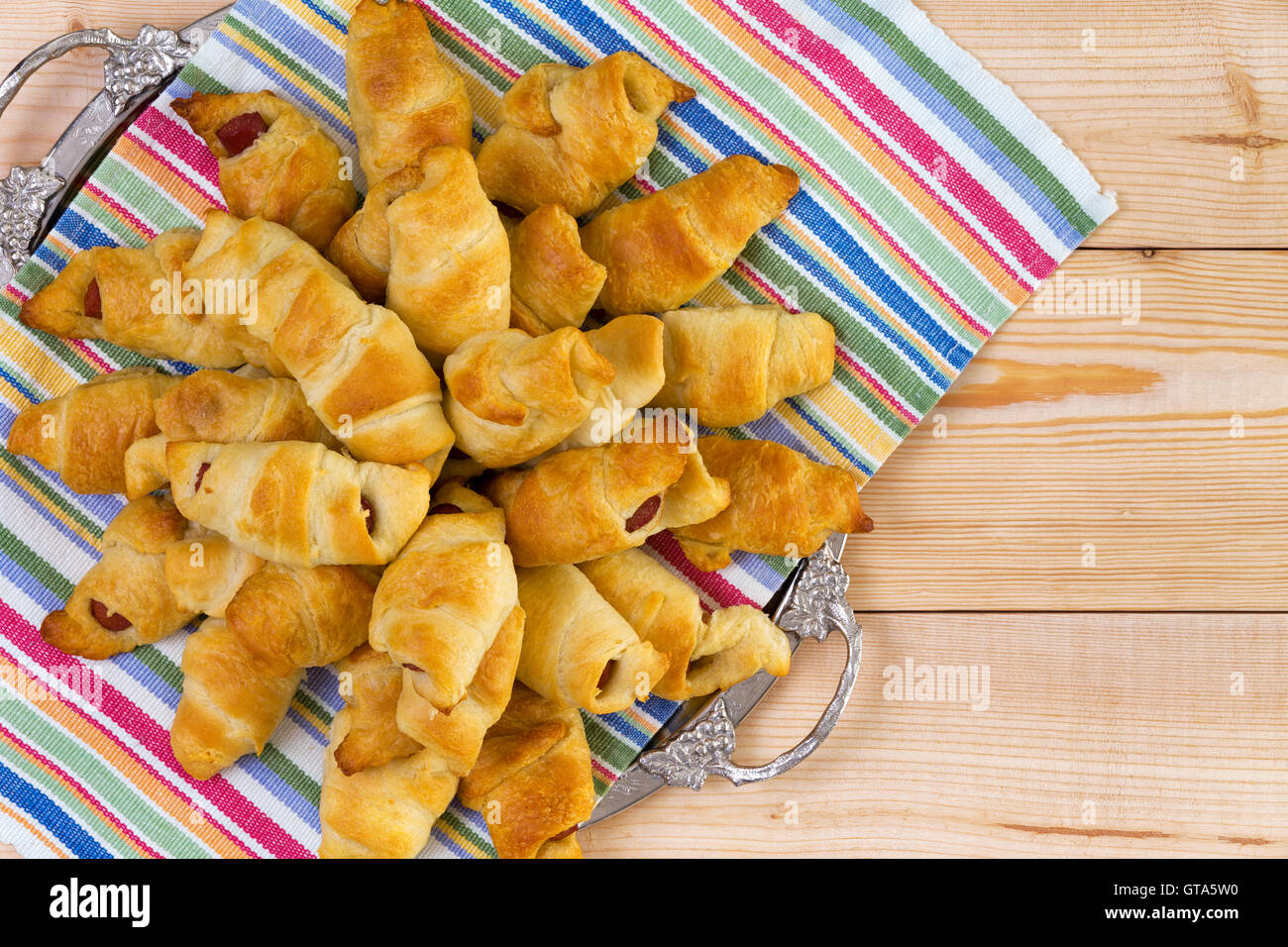 Haufen von köstlichen frischen Hot-Dog-Croissants auf einem bunten Serviette und Tablett auf einem hölzernen Picknicktisch für leckeres Finger Food summ Stockfoto
