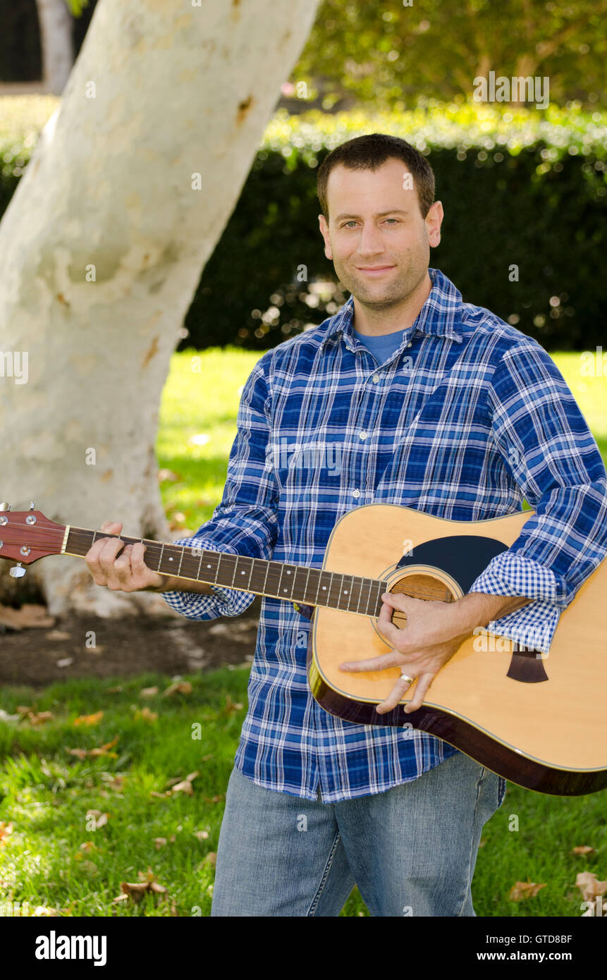 Mann spielt Gitarre in einem Park außerhalb trägt ein Hemd mit langen Ärmeln. Stockfoto