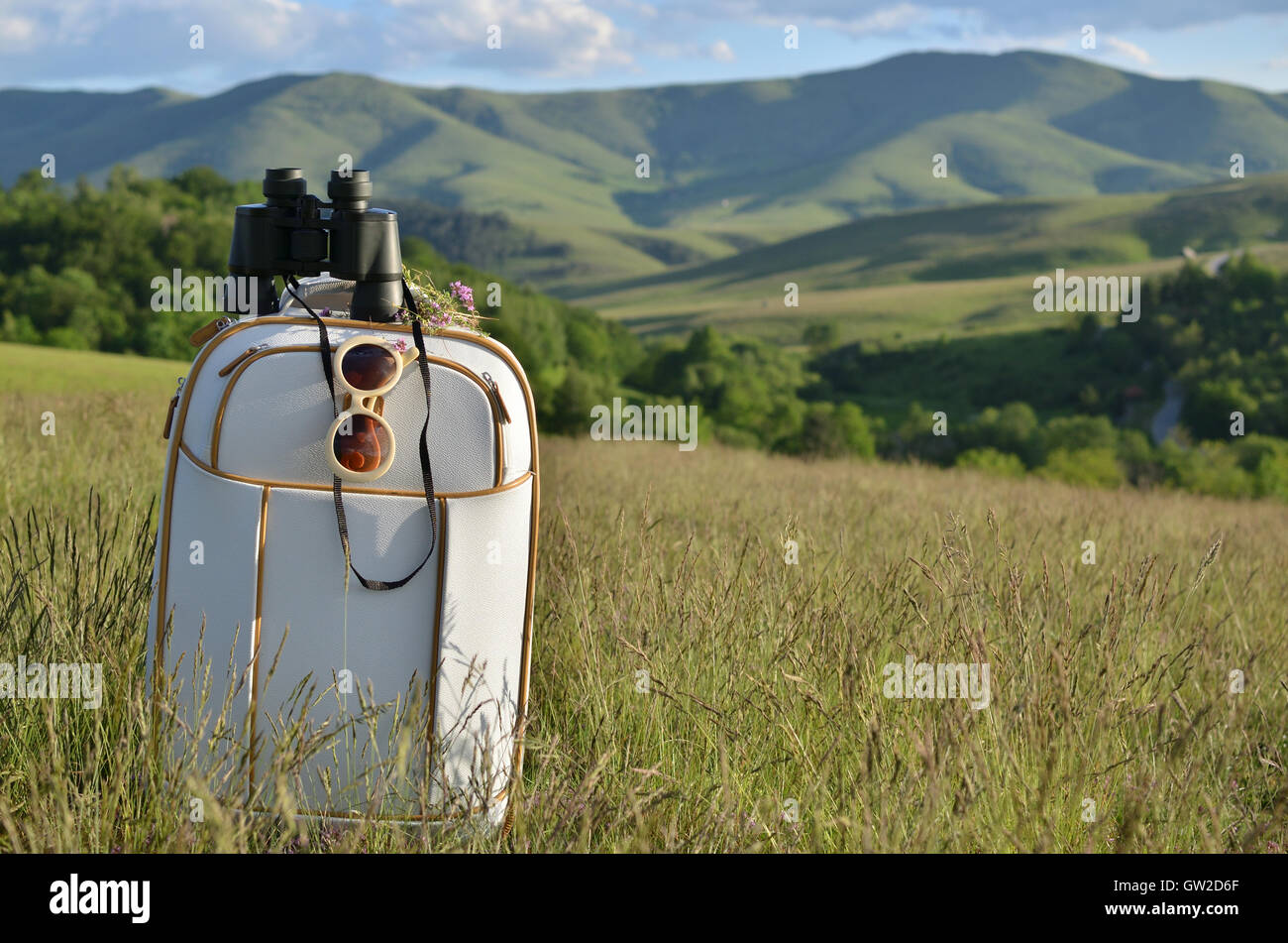 Weißen, eleganten Koffer mit Fernglas und Sonnenbrille auf der Bergwiese mit Hügeln und Himmel im Hintergrund Stockfoto