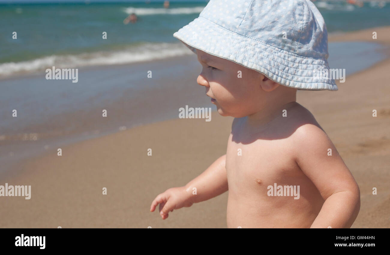 Glückliches Baby junge mit blauen Hut sitzt auf dem Sand und das Meer am Strand beobachten Stockfoto