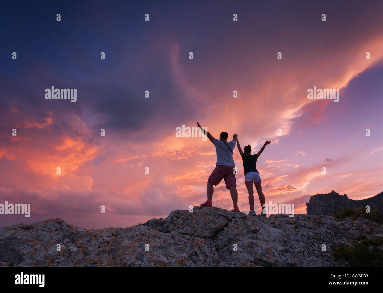 Landschaft mit Silhouetten von stehender junger Mann und Frau auf dem Berggipfel an farbenprächtigen Sonnenuntergang. Mann und Mädchen Stockfoto