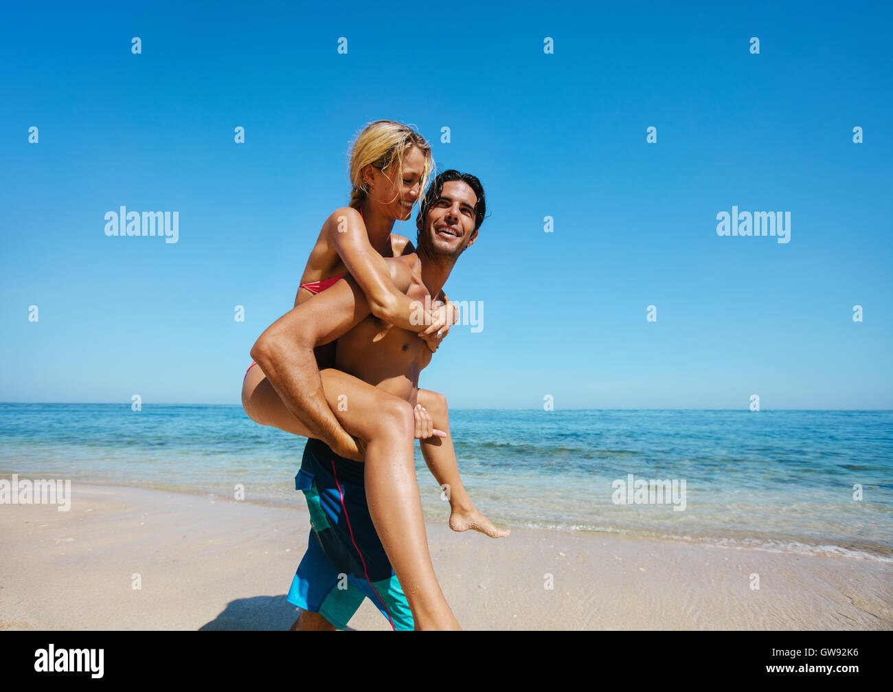 Richtungskontrolle Blick des jungen Mannes Pflege seiner Freundin auf zurück am Strand. Paar genießt Huckepack am Ufer Meeres. Stockfoto