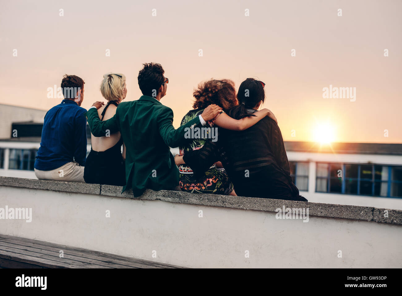 Rückansicht der jungen Männer und Frauen entspannen zusammen auf Dach und Blick auf den Sonnenuntergang. Gemischtrassig Freunde sitzen auf der Terrasse in e Stockfoto