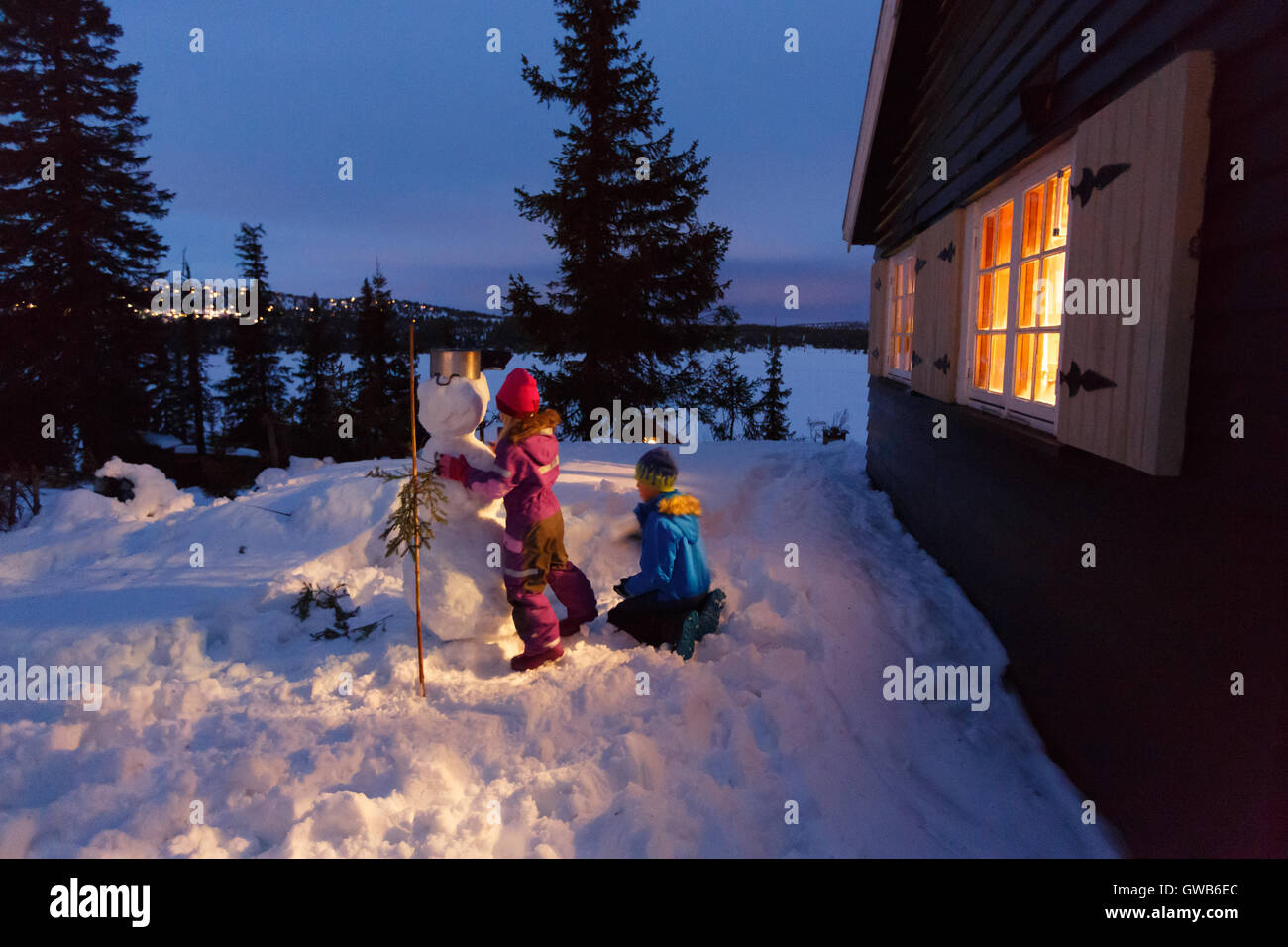 Seine New Years Eve und Kinder machen einen Schneemann. Die blaue Stunde draußen vor dem Fenster einer Winter-Hütte in den Bergen Norwegen Stockfoto