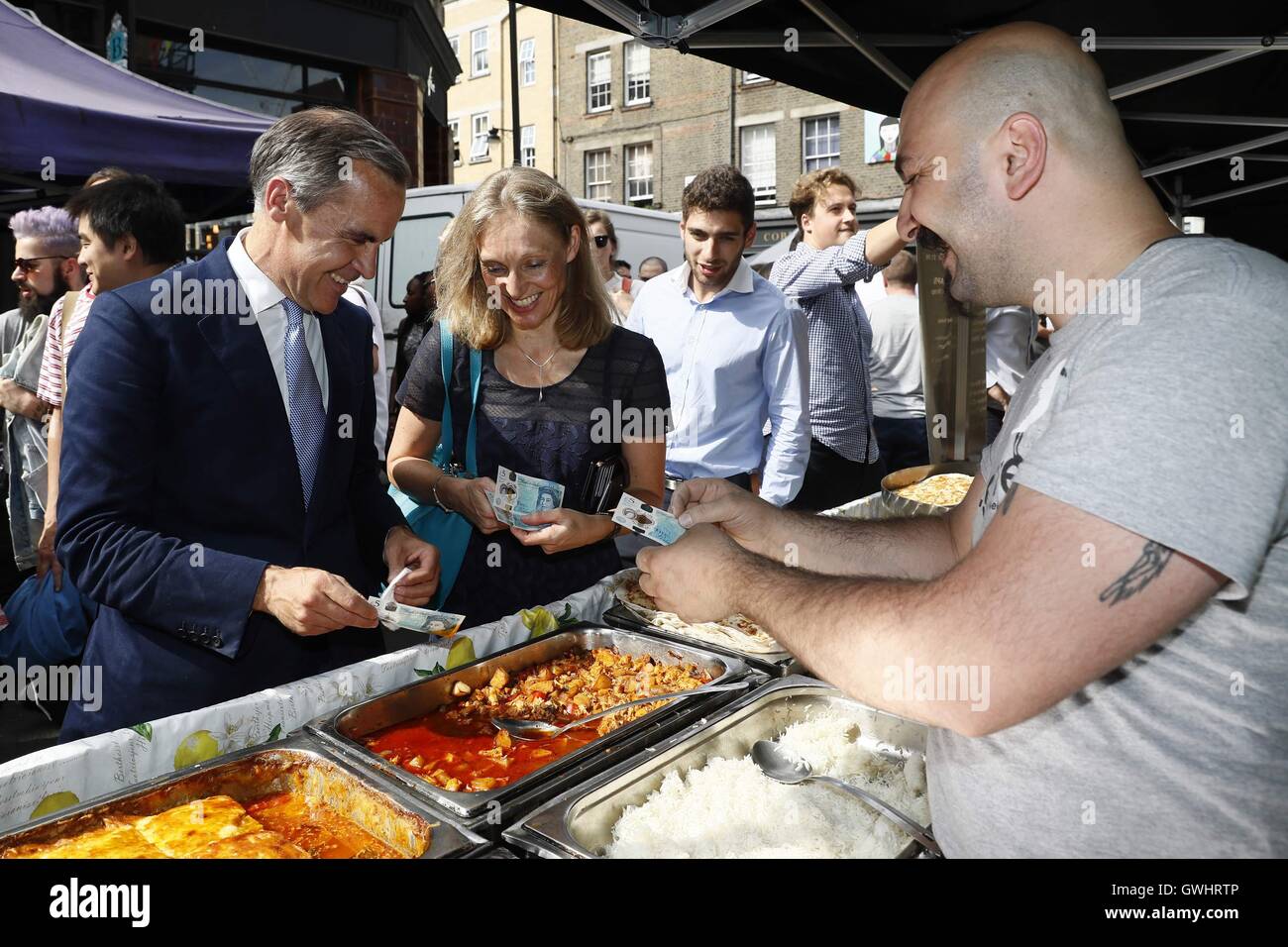 Bank of England Gouverneur Mark Carney (links) Dips eine neue Kunststoff £5 beachten Sie in ein Tablett mit Essen, wie er kauft Mittagessen aus einem Stall in Whitecross Street market in London. Stockfoto