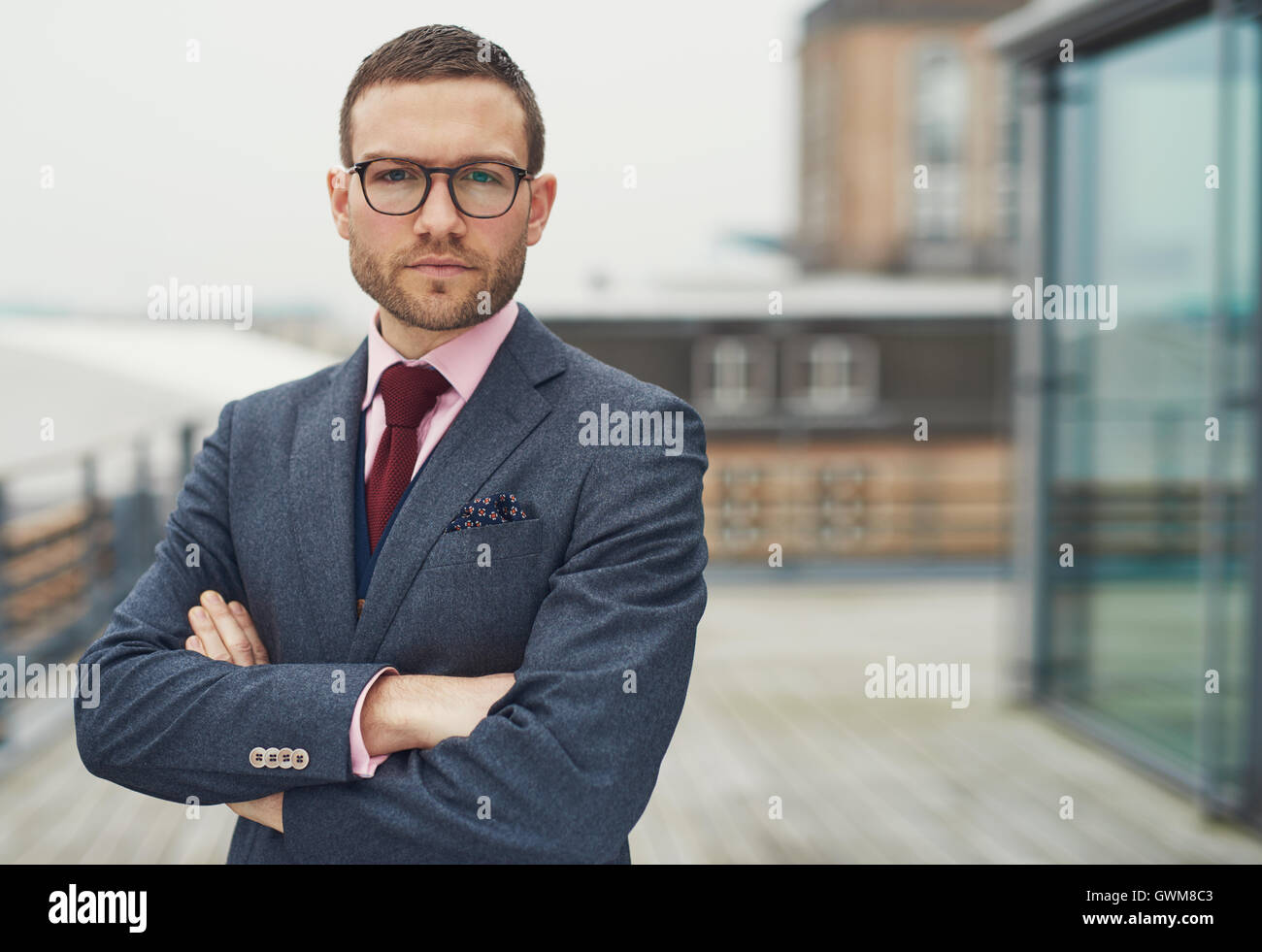 Zuversichtlich stilvollen Geschäftsmann mit Brille stehend auf einer open-air-Balkon schaut nachdenklich in die Kamera mit gefalteten arm Stockfoto