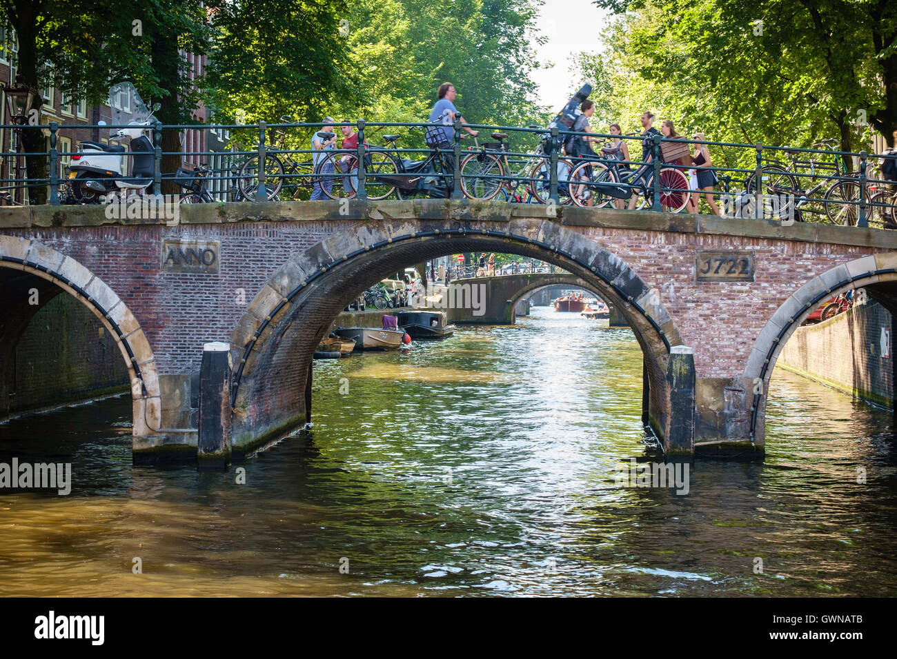 Radfahrer und Touristen überqueren eine sonnenbeschienene Kanalbrücke in Amsterdam. Stockfoto