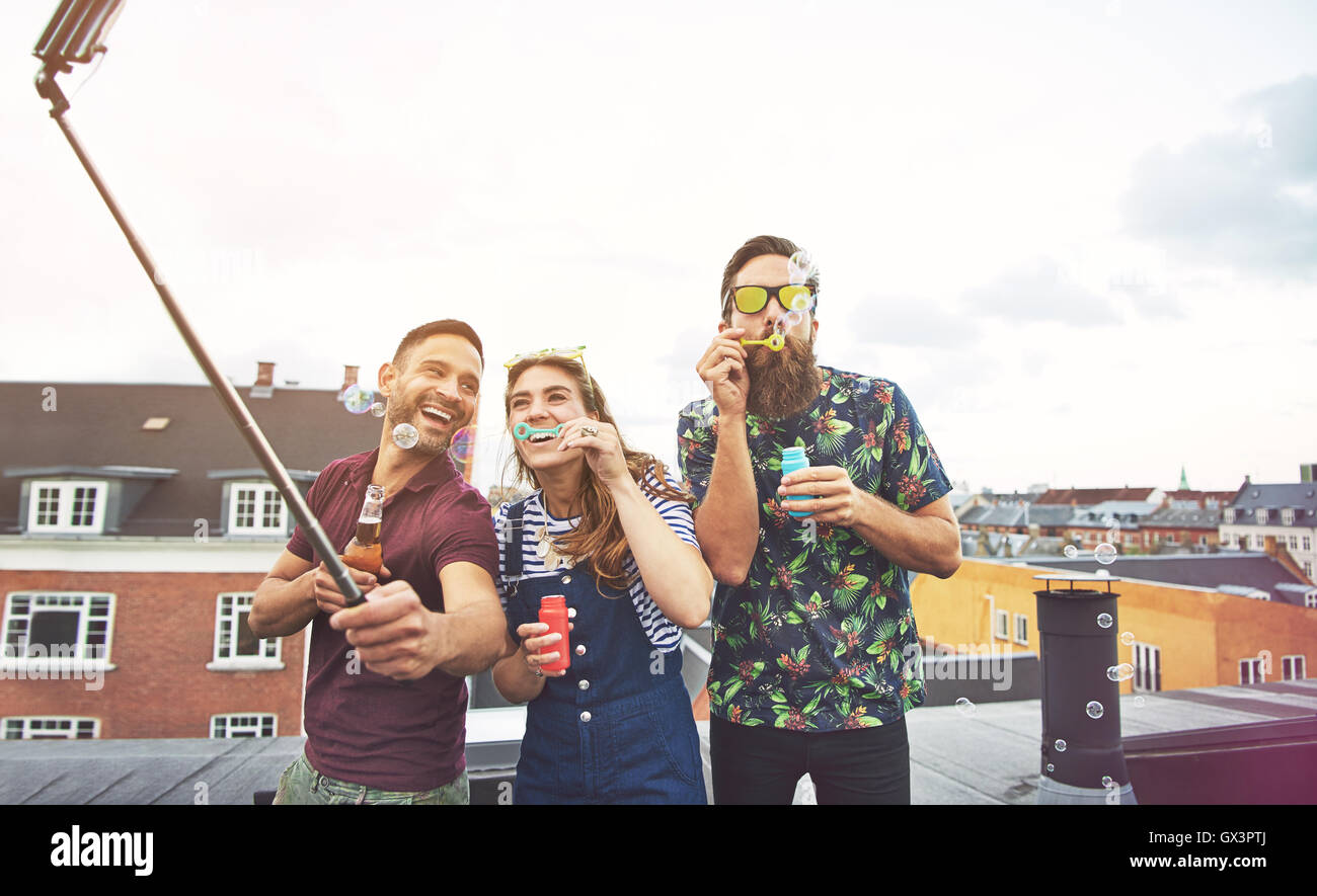 Drei Betrunkene Freunde, die Bilder von sich selbst auf dem Dach mit Kamera auf selfie Stick während Seifenblasen und Bier trinken. Stockfoto
