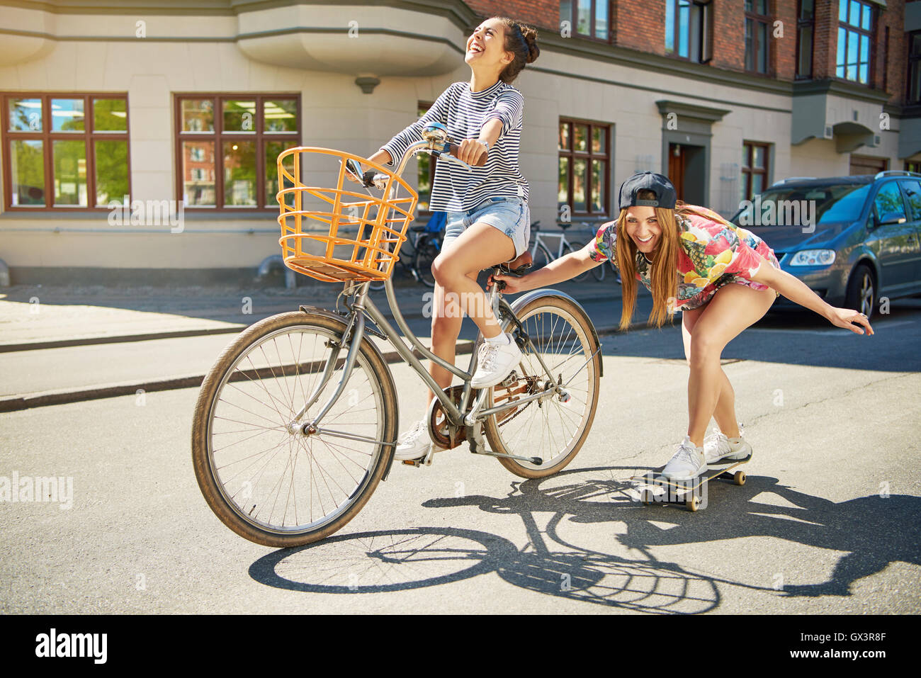 Lachend trendige junge Frau auf einem Skateboard entlang einer städtischen Straße abgeschleppt von ihrem Freund auf einem Fahrrad, Stockfoto