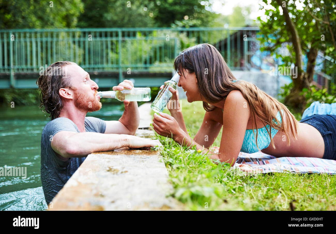 Junger Mann im Fluss, junge Frau liegt auf dem Rasen am Rand des Flusses, Mann aus Bierflasche trinken Stockfoto