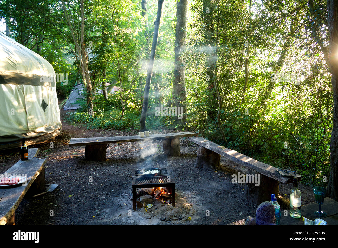 Lagerfeuer mit Rahmen, umgeben von 3 Holzbänke und eine Jurte Licht strömt durch Waldbäume, Zwiebeln Cooki Kochen Stockfoto