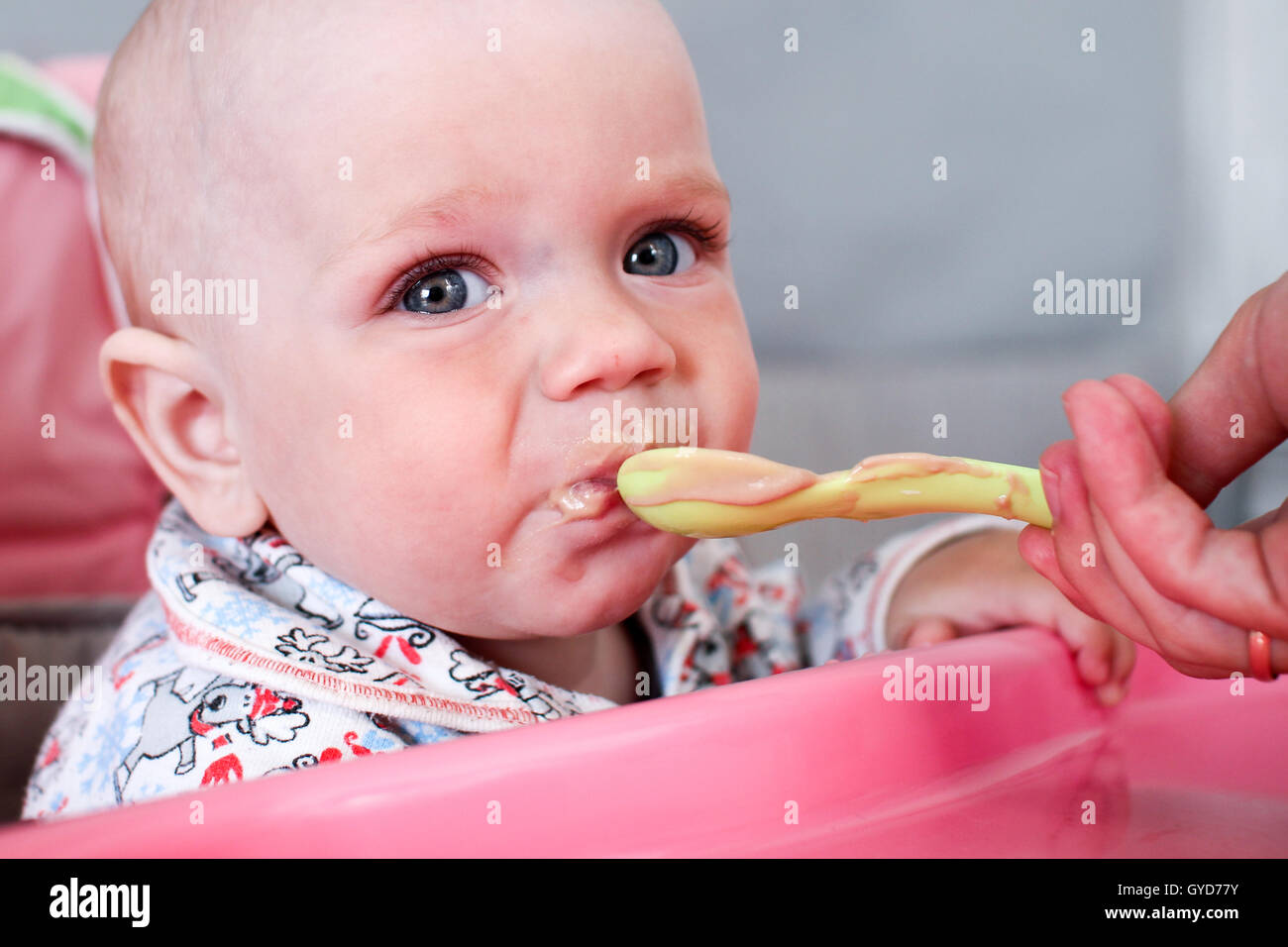 Schöne Baby isst Brei aus Mamas Hand. Er sitzt auf einem rosa Kinder "Stuhl. Stockfoto