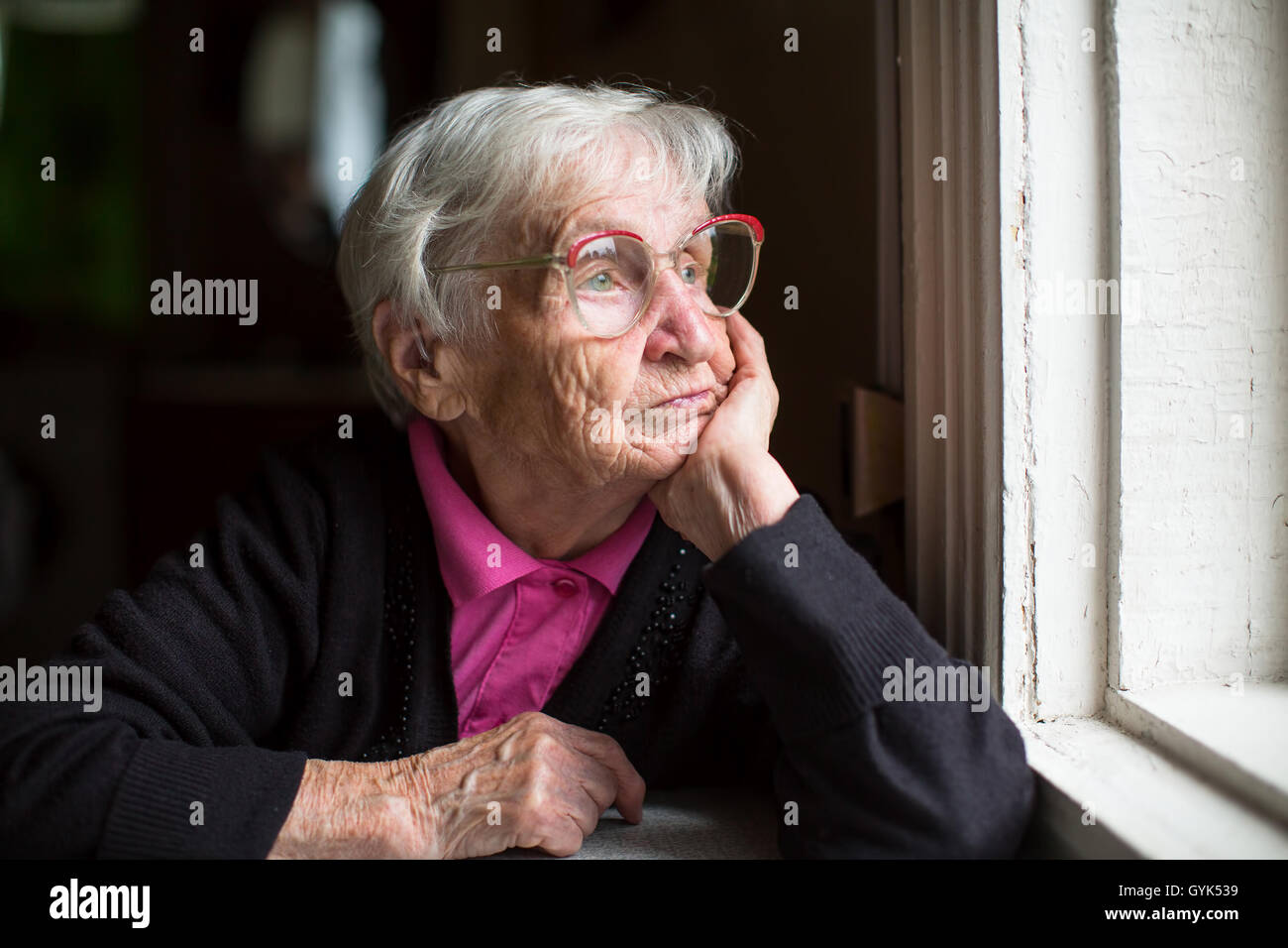 Ältere Frau in Gläsern nachdenklich schaut aus dem Fenster. Stockfoto