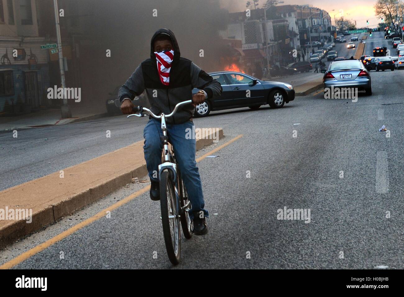 Ein Junge trägt ein Kopftuch über den Mund fährt Rad Weg von einem brennenden Auto während eines Aufstandes, Anschluss an die Beerdigung von Freddie Gray 27. April 2015 in Baltimore, Maryland.   Gray starb an den Verletzungen in Polizeigewahrsam. Stockfoto