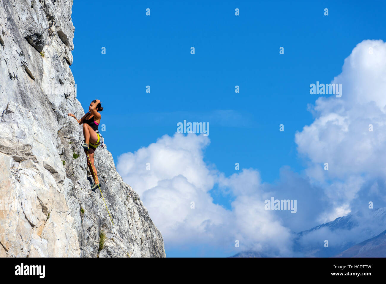 Gebräunte weibliche Kletterer in gelben Shorts auf steilen Felswand mit blauen Himmel und Wolken Stockfoto