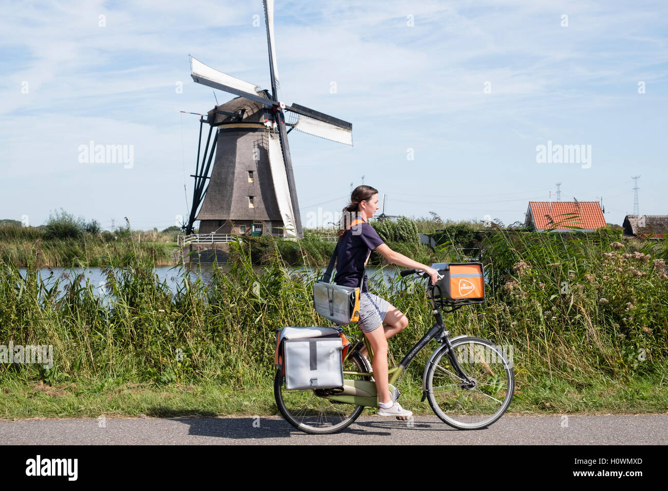 Junge Frau, die Zustellung mit dem Fahrrad für Postnl mit Windmühle bei Kinderdijk UNESCO World Heritage Site in The Hollands nach hinten Stockfoto