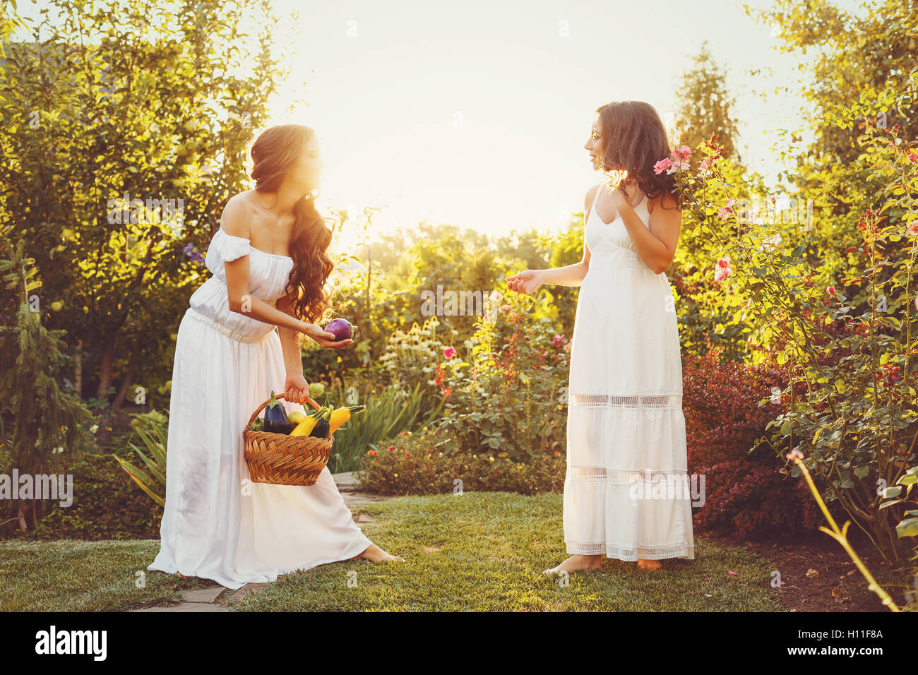 Zwei süße Schwestern in langen weißen Kleidern und einem Korb mit Ernte. Mädchen zeigt ihre Schwester Ernte. Reife Früchte und Gemüse. Stockfoto