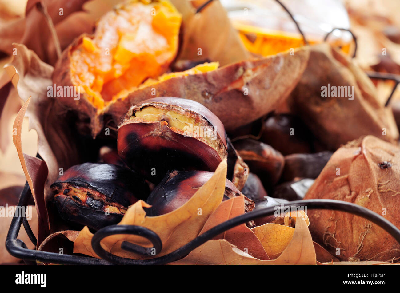 Einige geröstete Kastanien und einige geröstete Süßkartoffeln in einen Metallkorb mit Herbstlaub auf einem rustikalen Holztisch Stockfoto