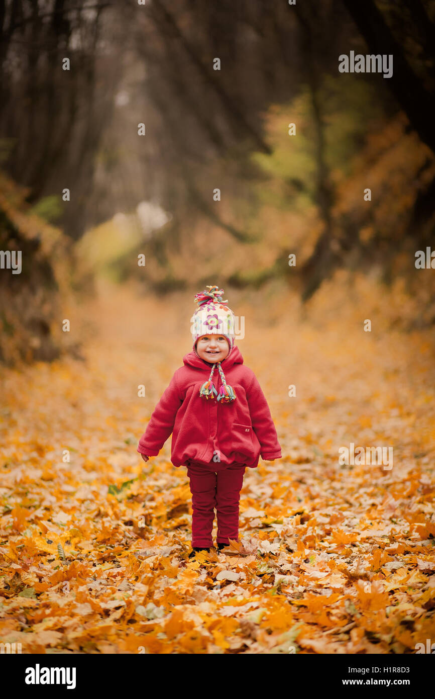 Kleines Mädchen im roten Mantel im malerischen Herbst Park. Baby mit gestrickter Mütze über Laub Hintergrund. Kleinkind Kind spielt mit gelb Stockfoto