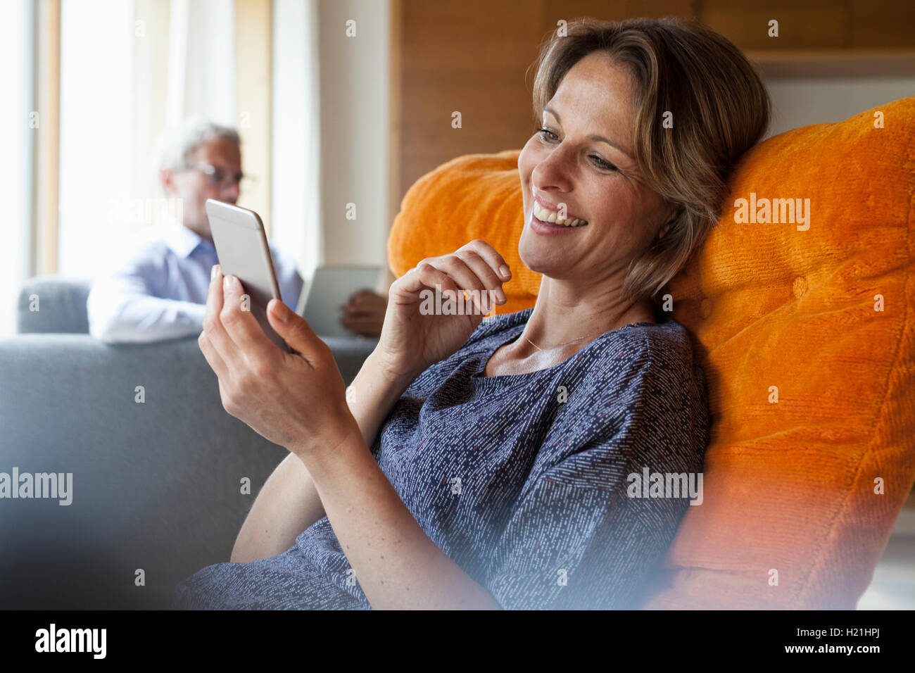 Lächelnde Frau zu Hause betrachten Handy mit Mann im Hintergrund Stockfoto