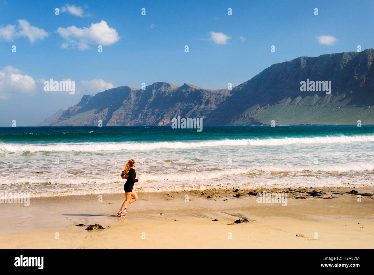 Junge Frau laufen Joggen am Strand Playa in La Caleta de Famara mit Klippen des Risco de Famara. Lanzarote, Kanarische Inseln Stockfoto
