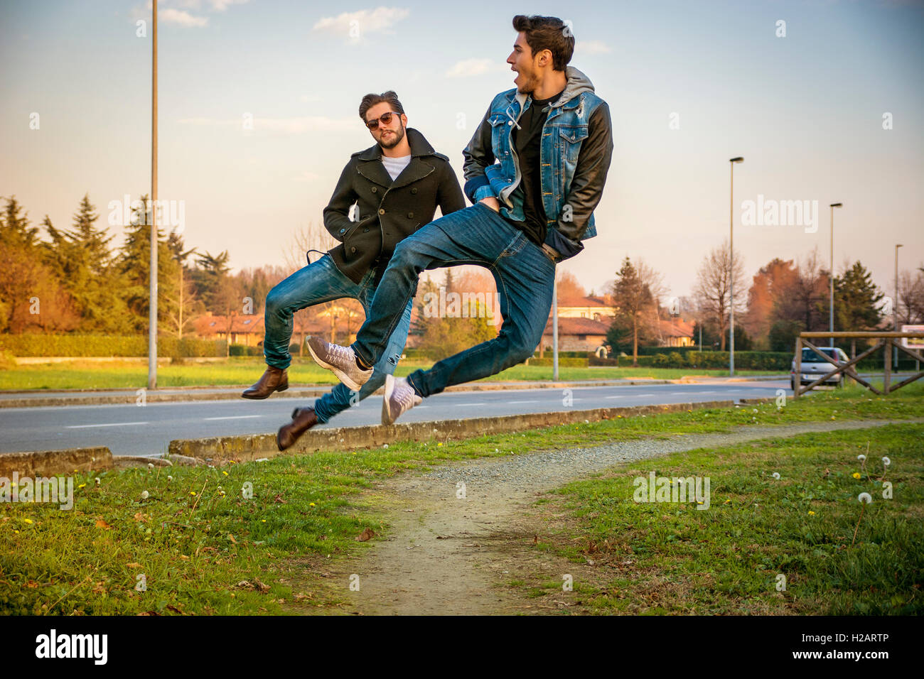Zwei junge Männer in die Luft springen und klicken Sie auf Fersen zusammen unisono während des Gehens auf Pfad im Stadtpark neben gepflasterten Roa Stockfoto