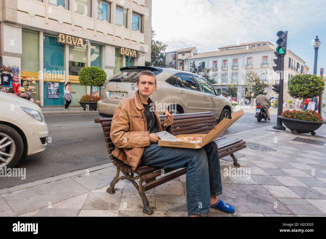 Junge Männchen Essen Pizza aus einer Kiste sitzend auf einer Bank der Stadt auf einem Bürgersteig mit Verkehr hinter ihm in Granada, Spanien. Stockfoto
