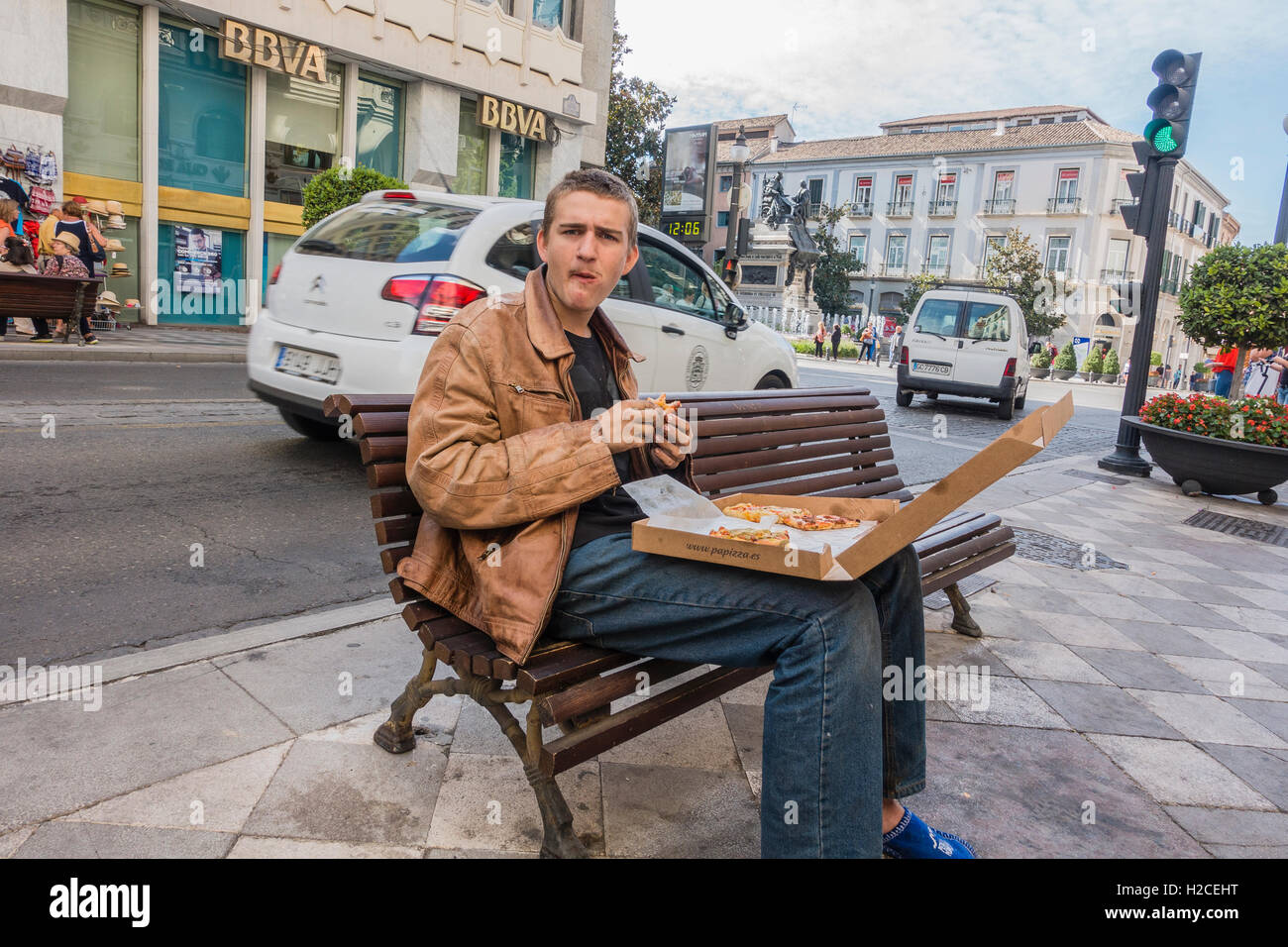 Junge Männchen Essen Pizza aus einer Kiste sitzend auf einer Bank der Stadt auf einem Bürgersteig mit Verkehr hinter ihm in Granada, Spanien. Stockfoto