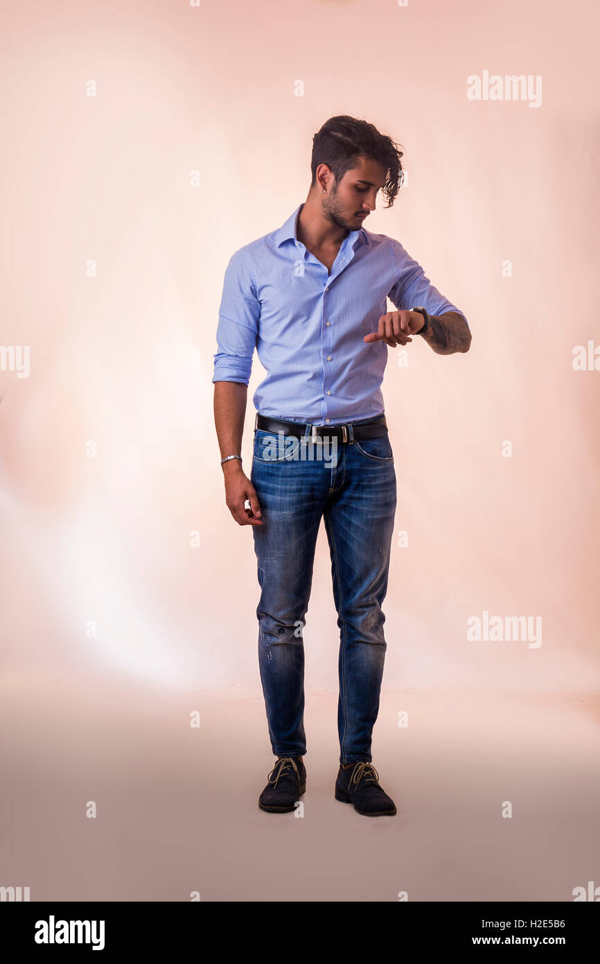 Porträt von Brünette junge Mann in hellblaues Hemd und Jeans, überprüfen der Zeit am Handgelenk beobachten, während in Studioaufnahme, steht Stockfoto